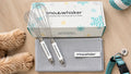 Snow Whisker dog grooming kit displayed on a light wood surface, including a white branded box, two metal wire whisks with white handles, a gray microfiber cloth, and a teal pet collar nearby and Goldendoodle paws. 